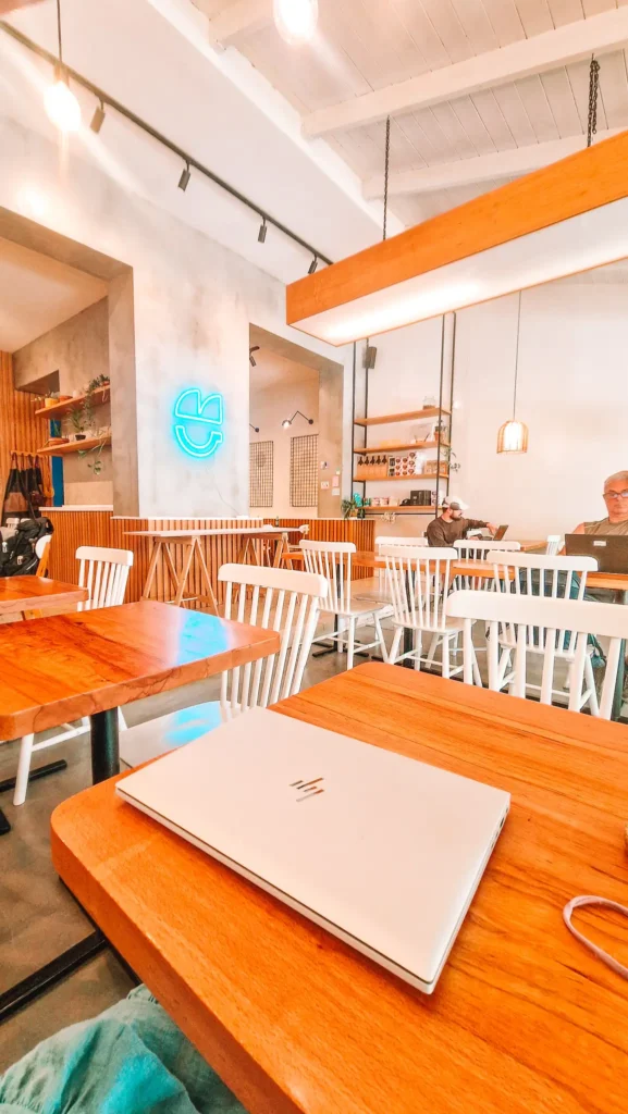 A laptop sits on a wooden table inside a bright modern café with white chairs and a turquoise neon logo. The spacious seating makes this café feel easy for remote work.